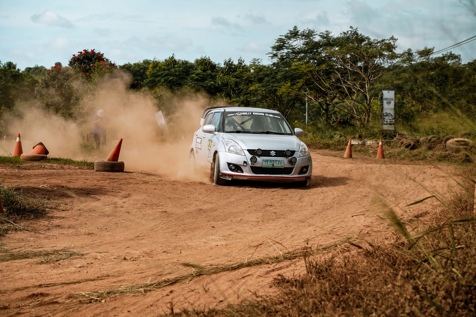 A rally car kicks up dust on a gravel stage lined with cones