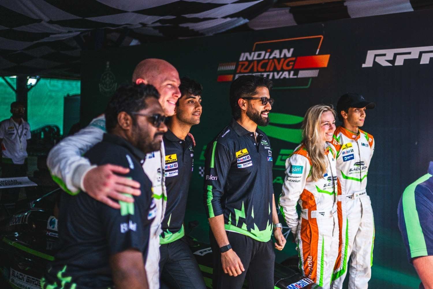 Drivers and team staff in racing suits pose under an Indian Racing Festival banner at a franchise team’s garage