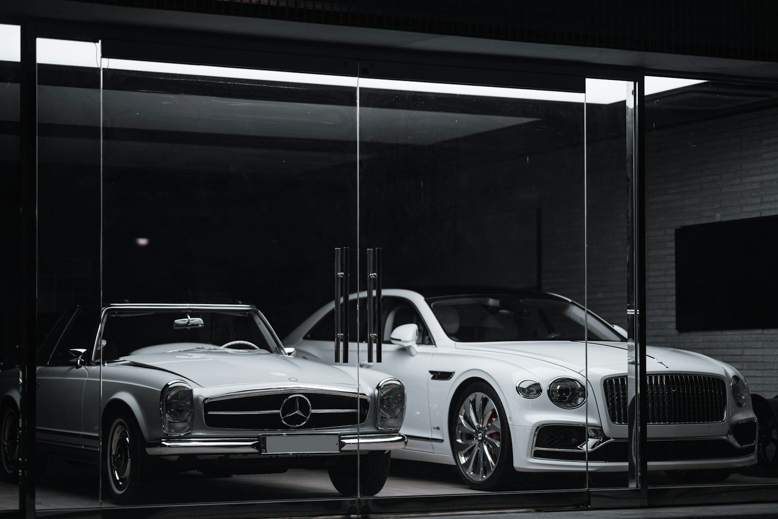 A vintage Mercedes-Benz convertible and a modern Bentley parked inside a glass-fronted luxury car showroom at night